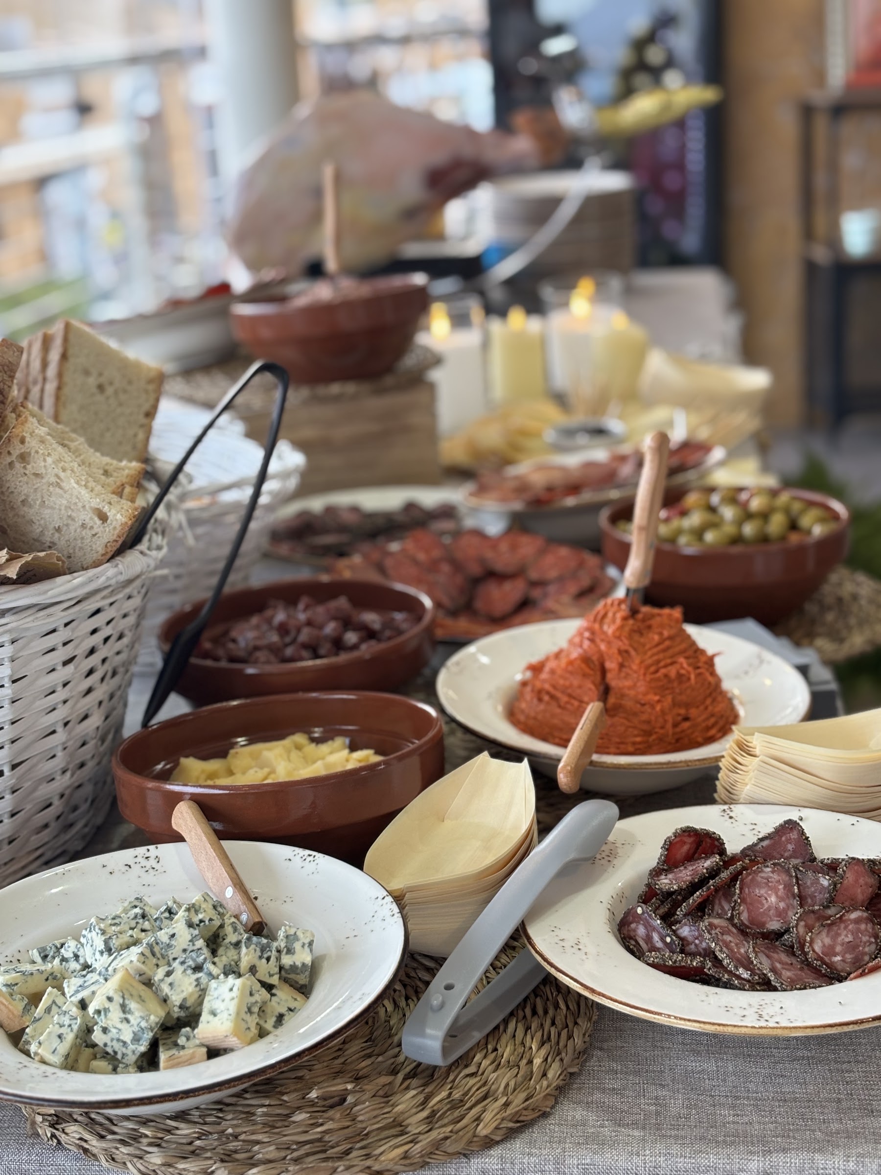 Mesa con quesos, salchichón, chorizo, aceitunas y pan, con jamón cortándose al fondo