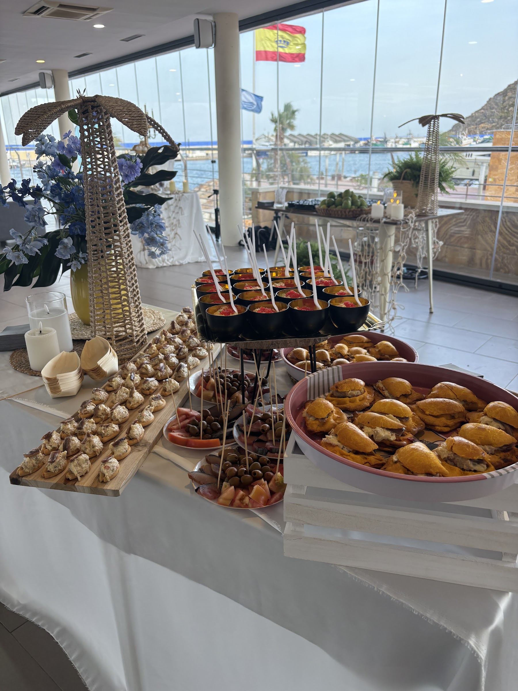 Mesa de evento montada en Mazmara Sunset con canapés y montaditos, y el puerto al otro lado del ventanal