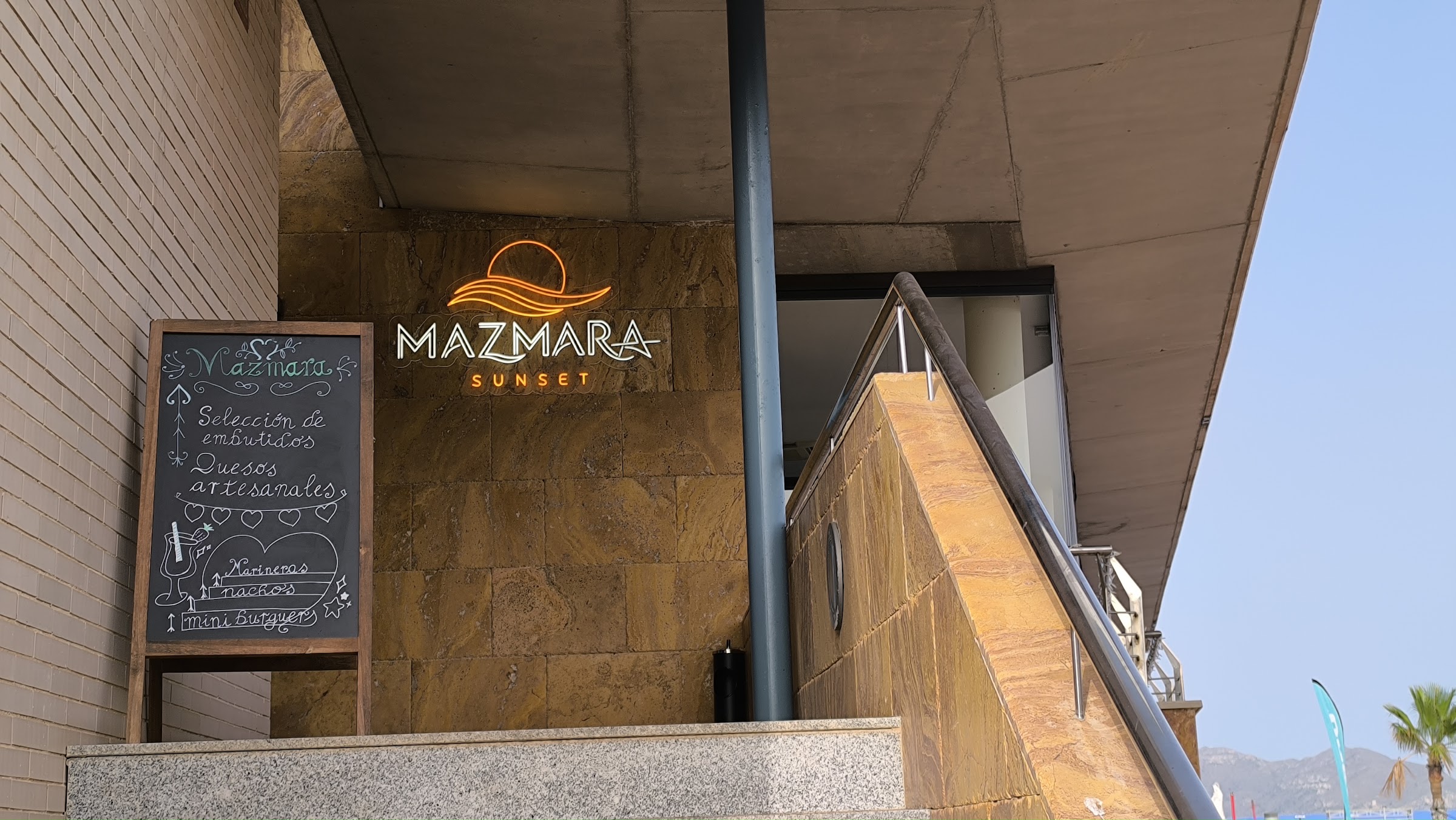 Entrada de Mazmara Sunset: el rótulo de neón con el sol sobre la ola y la pizarra con la carta del día
